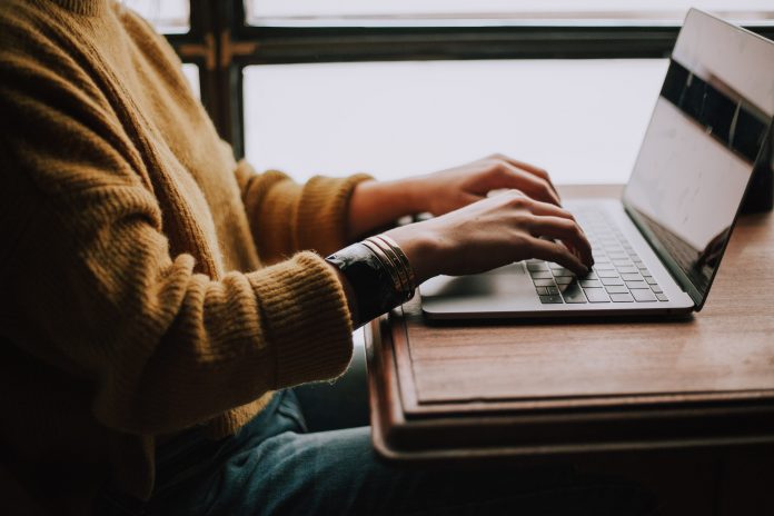 Woman working at a table, typing on her laptop. Only her arms and hands are seen. She is wearing a yellow sweater with the sleeves rolled up.