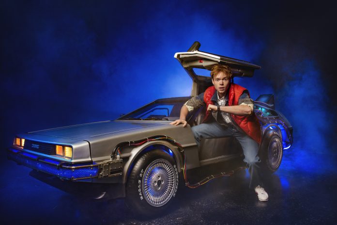 Ben Joyce as Marty McFly in Back To The Future The Musical. Joyce is climbing into a grey car and looking at the camera.