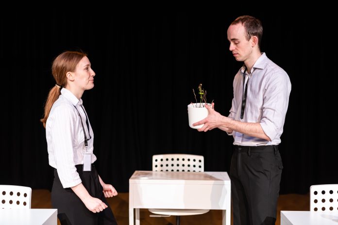 poster for slow violence. A man is looking down at a plant pot that he is holding. A woman smiles at him. They are in an office setting