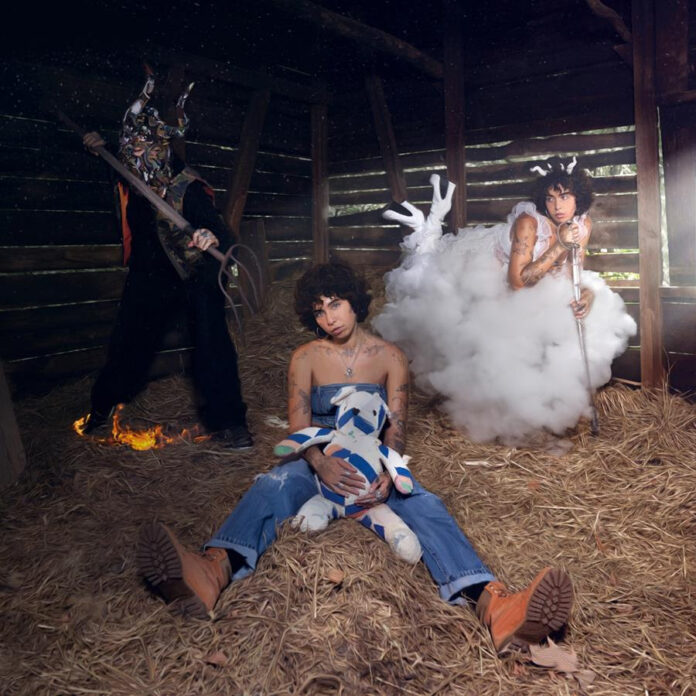 Album cover for Indigo De Souza's Wholesome Evil Fantasy. The artwork shows three figures in a dimly lit, rustic barn setting with straw on the floor. Indigo appears twice: once seated in the foreground wearing a strapless blue outfit, holding a patchwork plush toy with a tired expression, and once to the right, dressed in a cloud-like costume with white antlers, leaning on a staff. A third figure, in a dark, animalistic mask and wielding a large pitchfork, stands menacingly in the background, with their feet surrounded by small flames.