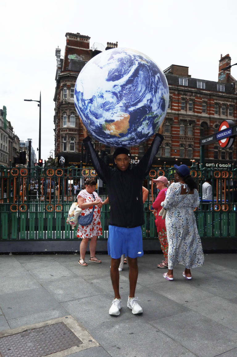 Rejjie Snow poses in front of city metro holding a globe in his hands above his head.